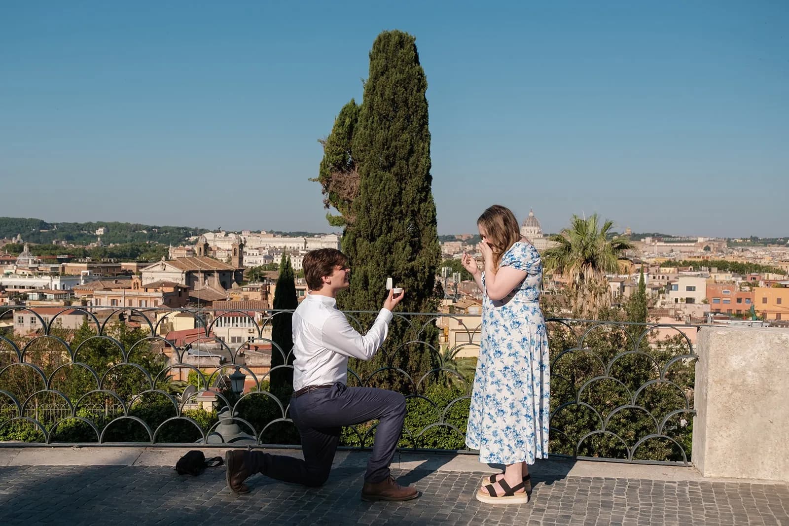 Gavin on one knee holding out the ring as Ally covers her face, on a terrace above the rooftops of Rome