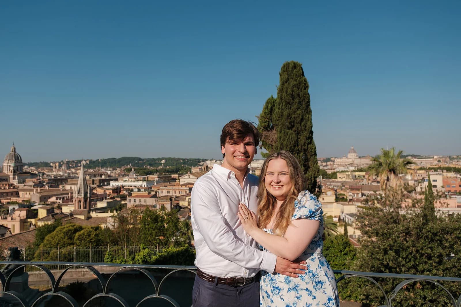 Gavin and Ally hugging and smiling on a terrace above the rooftops of Rome, Ally showing her engagement ring